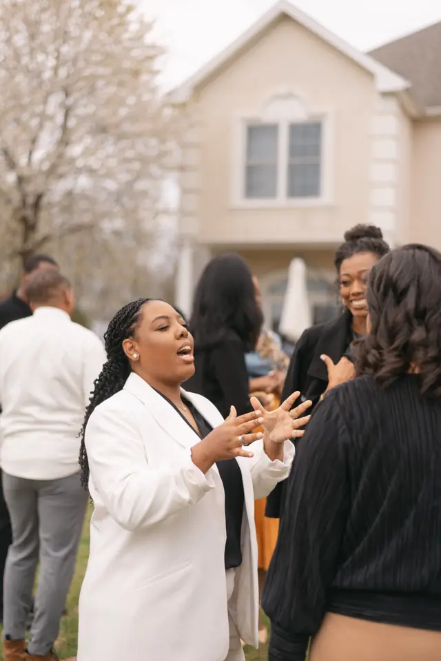 A woman in a white blazer speaks with her hands open while her sisters listen and smile.